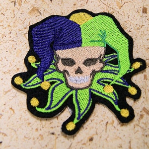 May include: Embroidered patch featuring a skull wearing a jester's hat. The hat is divided into blue, green, and yellow sections. The skull is beige with black eye sockets and a white-toothed grin. The patch has a black border and green and yellow accents.