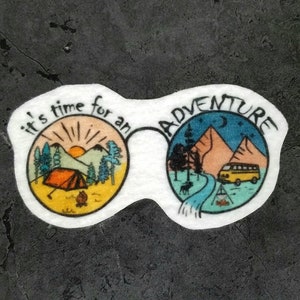 Peut inclure: Un autocollant blanc avec un motif de lunettes. La lentille gauche représente une scène de lever de soleil avec une tente, des arbres et des montagnes. La lentille droite représente une scène nocturne avec une camionnette, des arbres, des montagnes et une lune croissante. Le texte "it's time for an adventure" est écrit autour des lunettes.