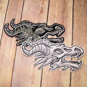 May include: Two embroidered iron-on patches featuring dragon skulls. The top patch is black and white with a detailed design. The bottom patch is white and gray with a more stylized design.