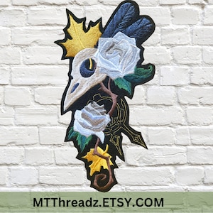 May include: Embroidered patch featuring a black and white skull with a bird beak, surrounded by white roses and gold leaves. The patch is on a white brick background.