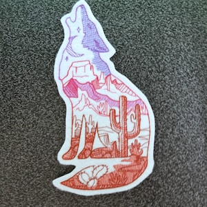 May include: An embroidered patch depicting a howling wolf with a desert landscape inside. The wolf is outlined in purple and the desert landscape is outlined in red. The desert landscape includes a cactus, mountains, and a crescent moon.