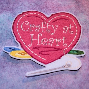 May include: Iron-on patch featuring an embroidered pink heart with the text "Crafty at Heart". The heart is outlined in white and has a white thread running through it. There are also three buttons and a needle and thread on the patch.