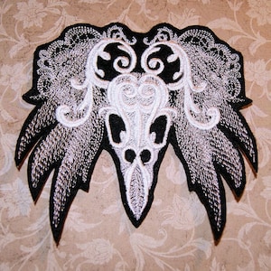 May include: Black and white embroidered patch with a skull design and intricate floral details. The skull is surrounded by a swirling pattern of leaves and vines.