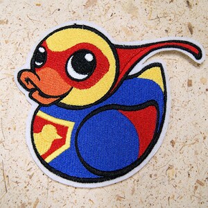 May include: Embroidered patch of a yellow, red, and blue superhero duck with a cape. The duck has large eyes and a determined expression.