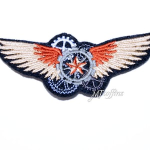 May include: A black embroidered patch with a beige and orange winged design. The wings are centered around a silver and black gear with a star in the center.