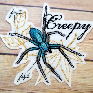 May include: Embroidered iron-on patch featuring a blue spider with black legs on a white background with gold leaves and the word "Creepy" in black cursive.
