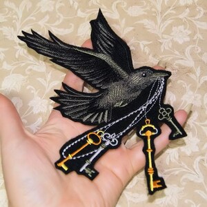 May include: Black embroidered iron-on patch of a crow carrying a chain of keys. The crow is in flight with its wings spread. The keys are gold and silver.