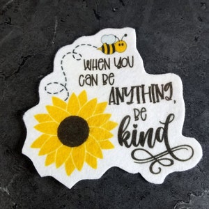 May include: White felt patch with a yellow sunflower and a bee flying above it. The text reads "When you can be anything, be kind."