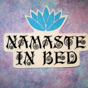 May include: White patch with black embroidered text "Namaste in Bed" and a blue embroidered lotus flower above the text.