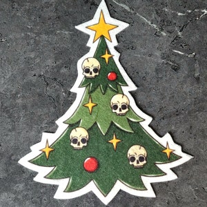 May include: A green felt Christmas tree with a yellow star on top. The tree is decorated with five white skulls and two red ornaments.