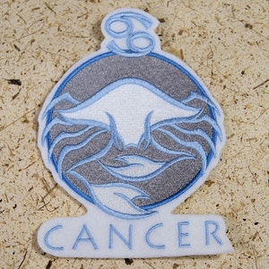 May include: A blue and grey embroidered iron-on patch featuring a stylised crab design and the word "CANCER" in white lettering.