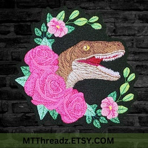 May include: Embroidered patch featuring a brown and white velociraptor head with an open mouth and sharp teeth, surrounded by pink roses and green leaves on a black background. The text "MTTThreadz.ETSY.COM" is at the bottom of the patch.