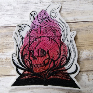 May include: Embroidered patch featuring a skull with a haunted house inside, surrounded by swirling smoke and ghosts. The skull is red and black, and the house is pink and black.