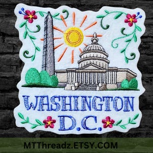 May include: Embroidered patch featuring a white background with a colorful depiction of the Washington Monument, the Capitol Building, and a sun with rays. The text "WASHINGTON D.C." is embroidered in blue below the buildings. The patch is surrounded by green vines and pink flowers.