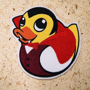 May include: A yellow rubber duck wearing a red cape and a burgundy tuxedo with a white bow tie. The duck has fangs and large eyes. The duck is a Halloween themed patch.