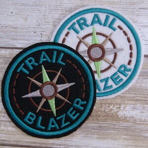 May include: Two embroidered patches with a compass design and the text "TRAIL BLAZER" in a turquoise blue circle. One patch has a black background and the other has a white background.