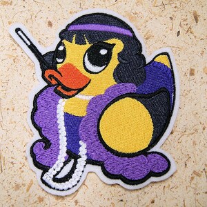 May include: A yellow duck with a purple and black outfit, a white pearl necklace, and a black wand. The duck has large black eyes and a red beak.