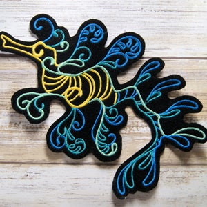 May include: Black embroidered patch in the shape of a seahorse. The seahorse design features yellow, teal, and blue details. The patch is made of a felt-like material and is likely an iron-on or sew-on applique.