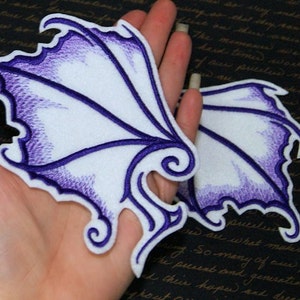 May include: Two white embroidered patches with purple detailing, shaped like wings. The wings have a jagged edge and a swirl design at the bottom.
