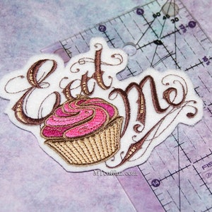 May include: Embroidered iron-on patch featuring the words "Eat Me" in brown cursive script and a pink and gold cupcake with swirl frosting.