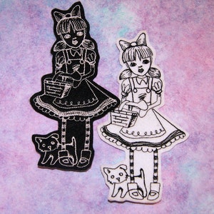 May include: Two black and white embroidered iron-on patches of girls with baskets and pets. The girl on the left is wearing a black dress and has a black cat. The girl on the right is wearing a white dress and has a white cat.