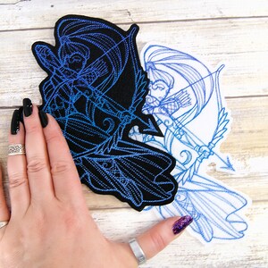 May include: Two embroidered patches featuring a blue outlined design of a woman with long hair holding a bow and arrow. The patches are on a white wooden surface. One patch is on a black background and the other is on a white background.