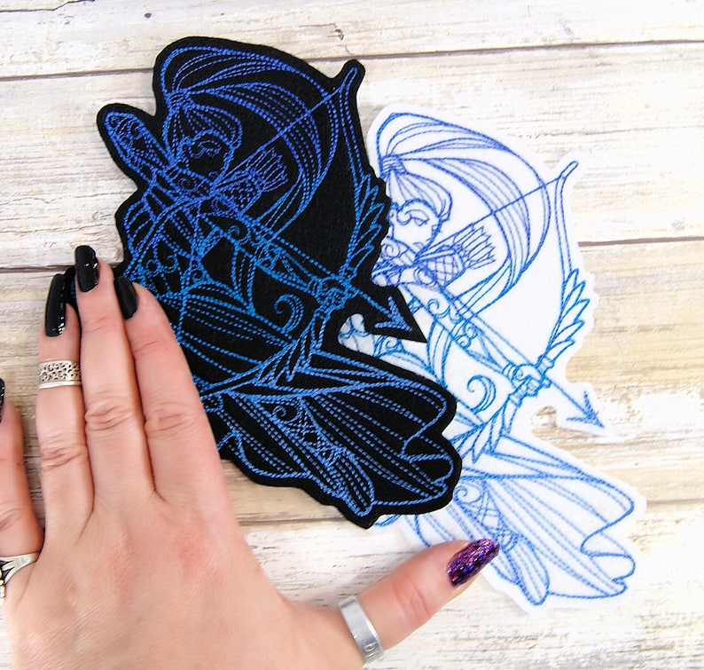 May include: Two embroidered patches featuring a blue outlined design of a woman with long hair holding a bow and arrow. The patches are on a white wooden surface. One patch is on a black background and the other is on a white background.