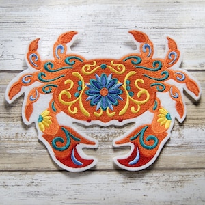 May include: Embroidered orange and blue crab with floral details. The crab is on a white background.