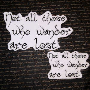 May include: Two white felt patches with black lettering that reads "Not all those who wander are lost". The patches are on a dark background.