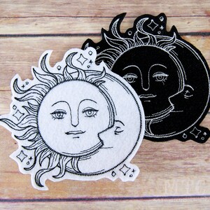 May include: Black and white embroidered patch featuring a sun and moon design. The sun has a face and rays, and the moon is a crescent shape with a face. The patch is on a wooden background.