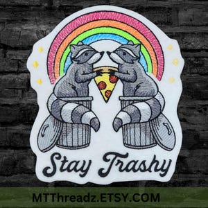 May include: Embroidered patch featuring two raccoons sharing a pizza slice under a rainbow. The raccoons sit atop trash cans, with the words "Stay Trashy" below. The patch has a white border and yellow star accents.