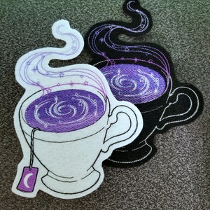 May include: Two embroidered teacups with purple and white swirling designs and a crescent moon tea bag tag. The teacups are layered on top of each other, with the top cup in white and the bottom cup in black.
