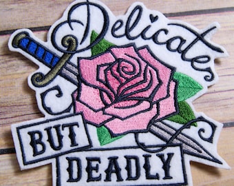 May include: Embroidered iron-on patch with a pink rose and a silver dagger, with the text "Delicates But Deadly" in black.