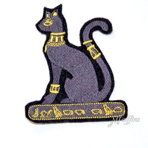 May include: An embroidered iron-on patch featuring a black cat with gold details. The cat is sitting with its tail curled around its body. The patch has a gold border with hieroglyphics.
