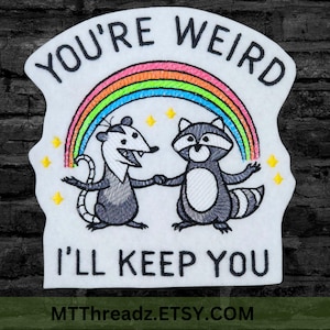 May include: Embroidered patch featuring a possum and raccoon holding hands under a rainbow. The text "YOU'RE WEIRD, I'LL KEEP YOU" is above and below the image. Yellow stars are scattered around the design.