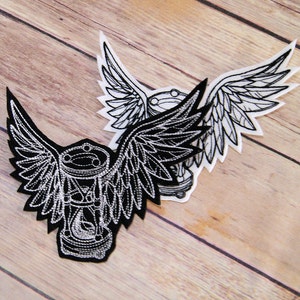 May include: Black and white embroidered patch of an hourglass with wings. The hourglass is surrounded by a circle of rings. The patch is on a wooden surface.