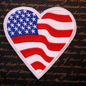 May include: A white heart-shaped patch with an embroidered American flag design. The flag is red, white, and blue with white stars on a blue field and red and white stripes.