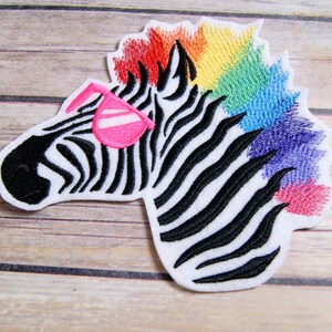 May include: Embroidered iron-on patch of a zebra with a rainbow mane wearing pink sunglasses. The zebra is facing left and has black and white stripes.