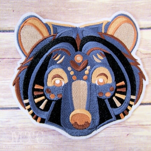 May include: Embroidered patch of a blue bear face with brown and tan details. The bear has a geometric design with a circular pattern around the eyes.