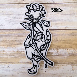 May include: White embroidered iron-on patch featuring a hand holding a rose with petals falling off. The text "White" is visible in the upper right corner of the image.