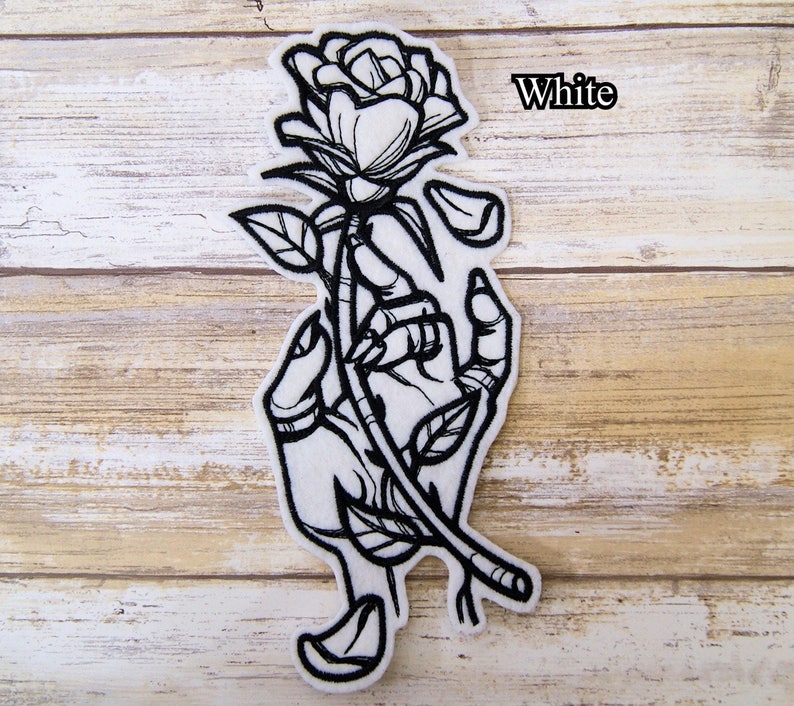 May include: White embroidered iron-on patch featuring a hand holding a rose with petals falling off. The text "White" is visible in the upper right corner of the image.