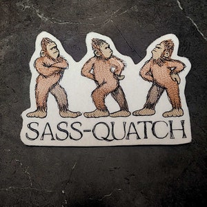 May include: A sticker featuring three cartoon Sasquatch figures in various poses, with arms crossed or hands on hips. The text "SASS-QUATCH" is printed in a serif font below the figures. The figures are brown with tan accents.