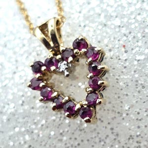 Può includere: Una collana con pendente a forma di cuore dorato. Il cuore è delineato da piccole pietre preziose rotonde, rosso scuro, con un'unica piccola gemma trasparente al centro. Anche la catena della collana è dorata.
