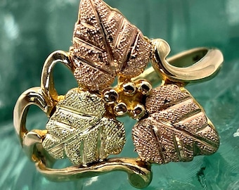 May include: A gold ring with a leaf and berry design. The ring features three leaves in shades of gold and rose gold, with small gold berries in the centre. The band and stems are gold.