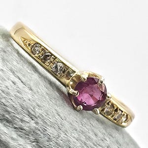 May include: A gold ring featuring a round, deep pink gemstone set in the center with four prongs. The band is accented with three small, clear, round-cut stones embedded along each side of the central setting, creating a symmetrical design.
