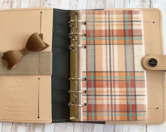 May include: Open leather Filofax personal organizer in beige with a brown bow and elastic band. The interior features a plaid patterned insert in shades of orange, teal, and cream. The organizer is stamped with "Filofax" and "Made in the UK".
