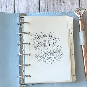 May include: A light blue Filofax planner with a silver binder, open to a page with a black and white illustration of a book, moons, and flowers. A rose gold pen with a diamond-shaped top rests beside the planner.