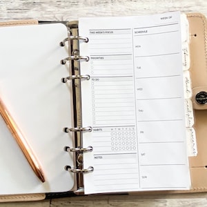 May include: A beige leather ring-bound planner featuring a weekly schedule insert with sections for focus, priorities, to-do lists, habits, and notes. A rose gold pen with a diamond-shaped topper rests on a blank white page inside the planner.