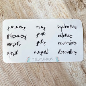 Puede incluir: Una hoja de pegatinas blanca con los meses del año escritos en cursiva negra. Los meses están dispuestos en tres columnas. En la parte inferior de la pegatina hay una pequeña ilustración de un unicornio y el texto "THELUCKYUNICORN".