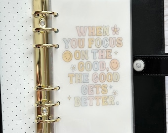 May include: A black binder with gold rings and a clear pocket insert. The insert features the text "WHEN YOU FOCUS ON THE GOOD, THE GOOD GETS BETTER" in pastel colours with smiley faces and flower graphics. The binder is open, revealing a polka dot interior.